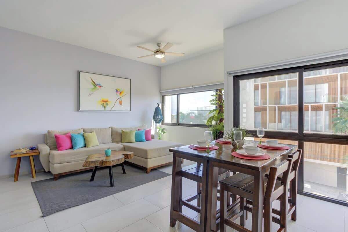 A modern living room with a sofa, coffee table, dining table, and chairs. There is a ceiling fan, a window with a view, and a painting on the wall.
