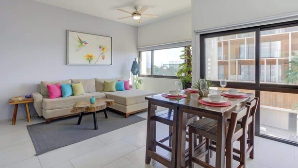 A modern living room with a sofa, coffee table, dining table, and chairs. There is a ceiling fan, a window with a view, and a painting on the wall. - Family oasis near the sea - La Cruz Encantada 3A (La Cruz de Huanacaxtle)
