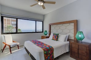 Ein Schlafzimmer mit einem Bett, einem Sessel, einem Nachttisch, einer Lampe und Blick auf das Meer durch ein Fenster.