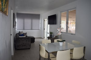 A modern living room with a sofa, a dining table with chairs, a TV mounted on the wall, and a window with blinds. - Villa Aerio (Carolina)