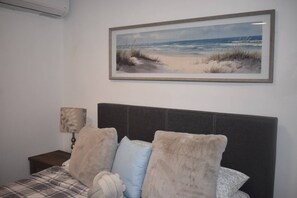 A bedroom with a bed, a nightstand, a lamp, and a framed painting of a beach scene. - Villa Aerio (Carolina)