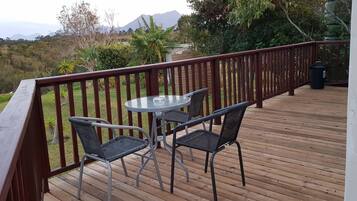 A wooden deck with a glass table and two chairs, overlooking a mountainous landscape.