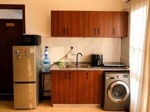 Eine kompakte Küche mit Holzausstattung, einem Edelstahlkühlschrank, einer Mikrowelle, einem Spülbecken, einer Kaffeemaschine und einer Waschmaschine.