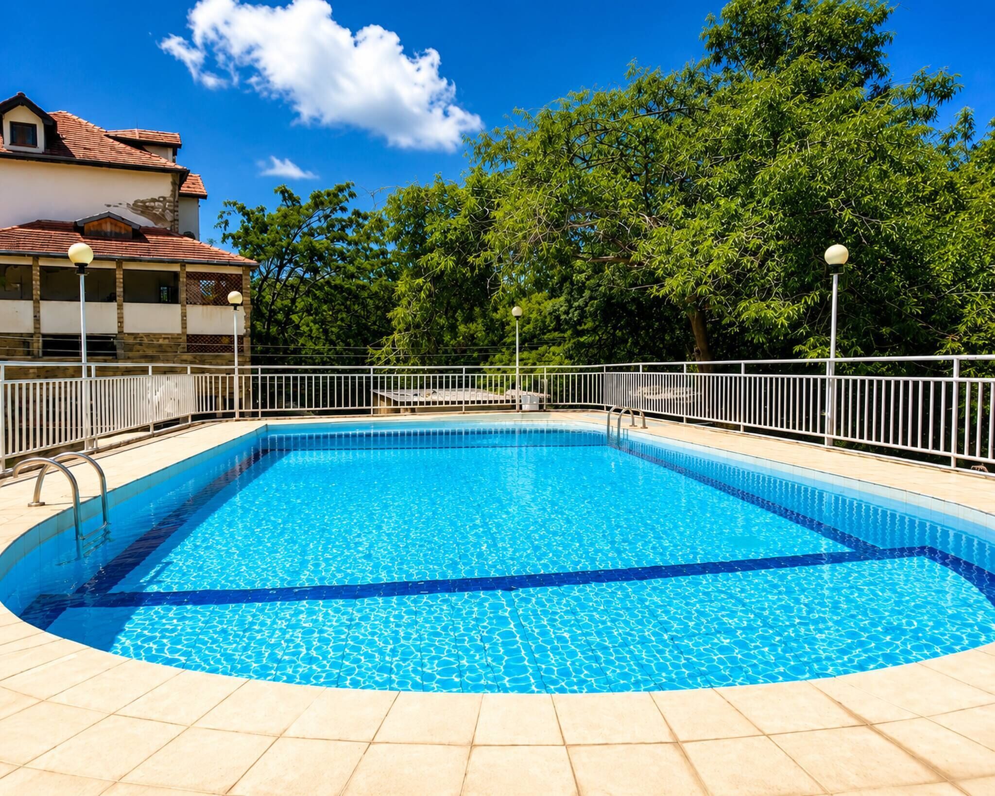 Ein rechteckiger Swimmingpool mit Fliesen umrandet, umgeben von einem weißen Metallzaun, mit Bäumen im Hintergrund.