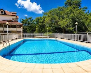Ein rechteckiger Swimmingpool mit Fliesen umrandet, umgeben von einem weißen Metallzaun, mit Bäumen im Hintergrund.
