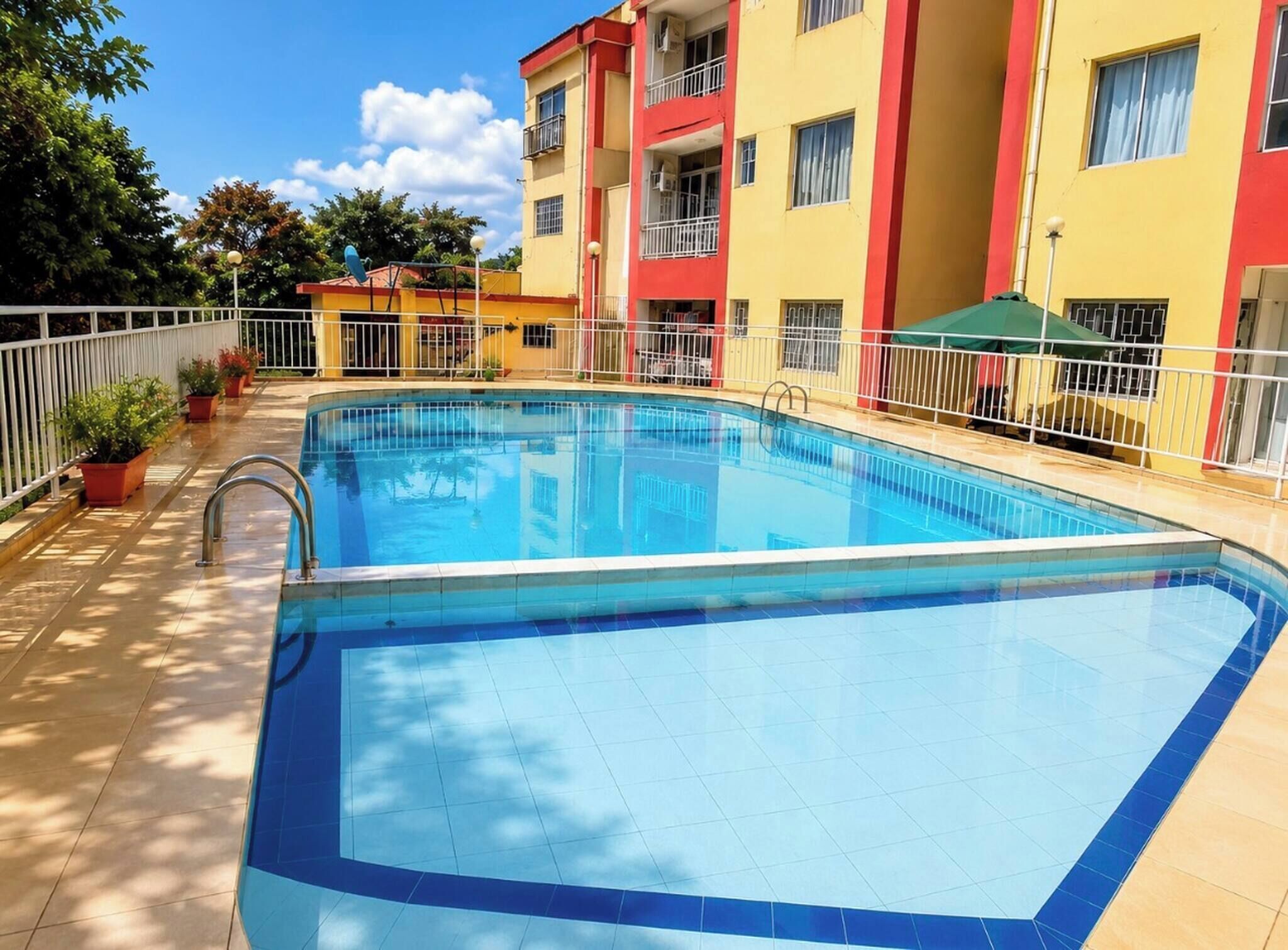 Una piscina con suelo de baldosas, rodeada por una cerca blanca, y edificios adyacentes con balcones.