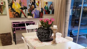 Et spiseområde med et bord dækket til fire personer, omgivet af hvide stole med sorte hynder. En sort vase med lyserøde blomster og en lysestage står på bordet.