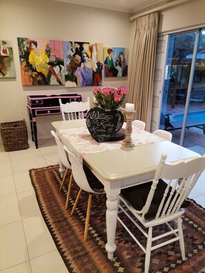 Un coin repas avec une table dressée pour quatre personnes, entourée de chaises blanches dotées de coussins noirs. Un vase noir contenant des fleurs roses et un chandelier sont posés sur la table.
