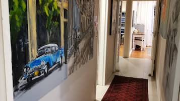 Een gang met een rood tapijt met patroon, een schilderij van een blauwe auto, en een deur die leidt naar een kamer met een witte stoel.