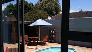 En swimmingpool med fliseterrasse, omgivet af en belagt terrasse. Træloungestole og et bord står under en hvid parasol.