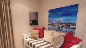 En hvid sofa med stribede puder og røde hovedpuder, et maleri af en havneudsigt, og et vindue med cremefarvede gardiner.