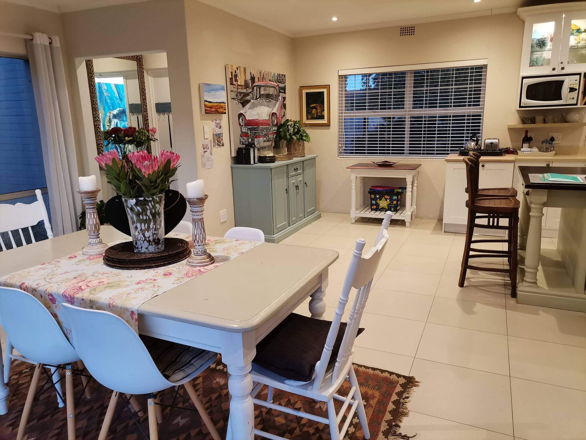 A dining area with a table set for four, chairs, a cabinet with decorative items, a window with blinds, and a kitchen area with a microwave.