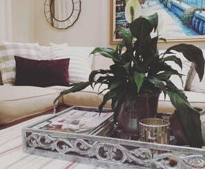 Sala de estar com sofá, planta em vaso, bandeja com revistas e um espelho decorativo.
