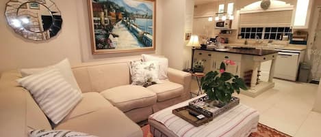 A living room with a sofa, a coffee table, a painting, and a kitchen in the background.