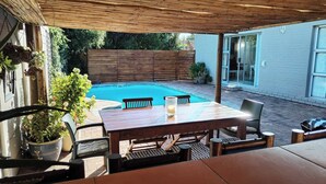 En udendørs patio med pool, træbord, stole og krukker med planter.