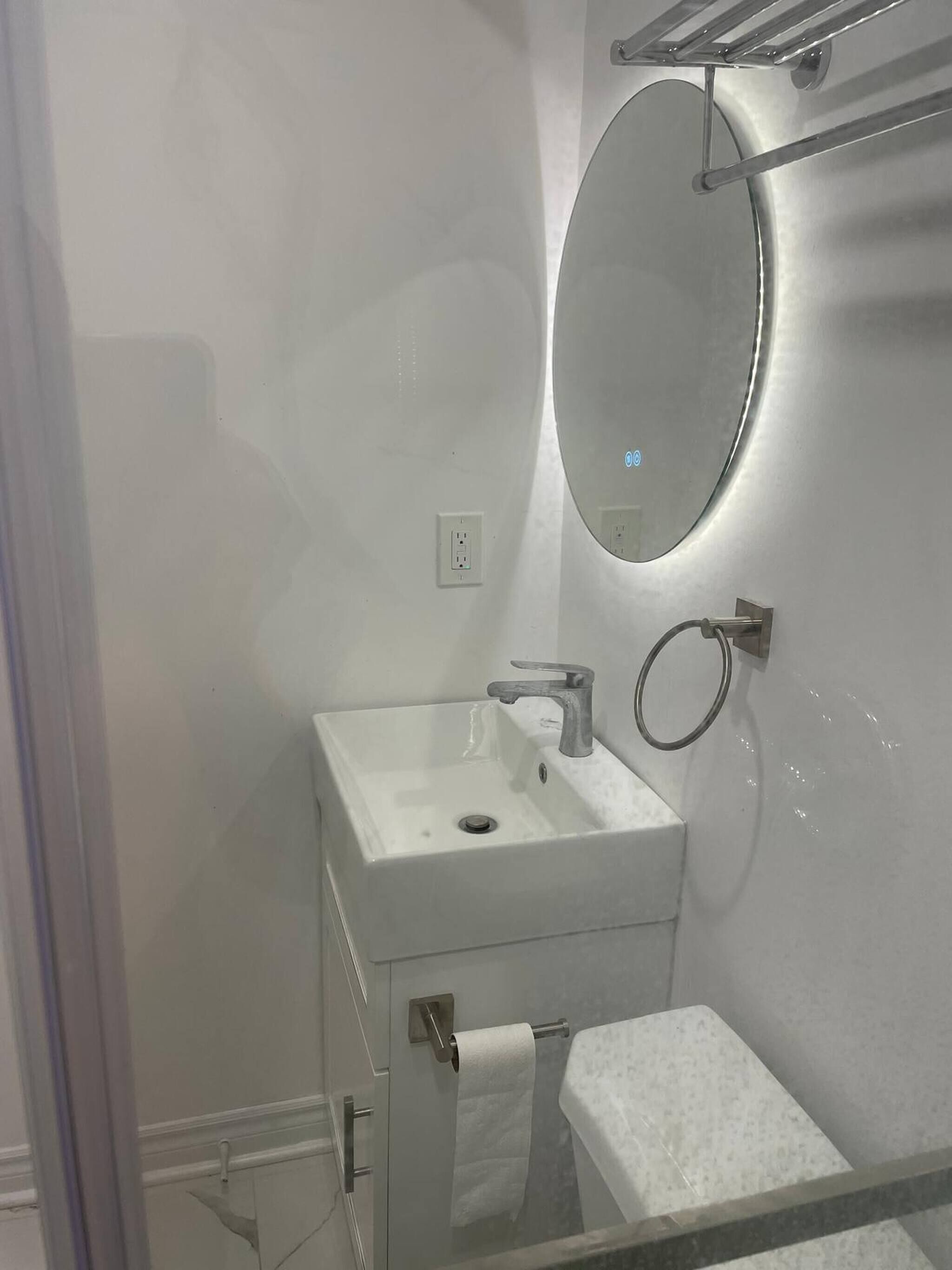 Un baño con un lavamanos blanco, un espejo, un perchero para toallas y una cabina de ducha con mampara de vidrio.