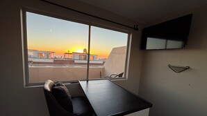 Huoneessa on työpöytä, tuoli, seinälle asennettu televisio ja ikkuna, josta avautuu näkymä kaupungin siluettiin auringonlaskun aikaan.