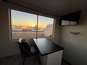 Ett rum med ett skrivbord, en stol, en tv som är monterad på väggen och ett fönster med utsikt över en stadssiluett i solnedgången.