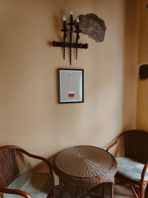 Una habitación con una mesa redonda de mimbre, dos sillas de mimbre, un cuadro enmarcado y un candelabro fijado a la pared.