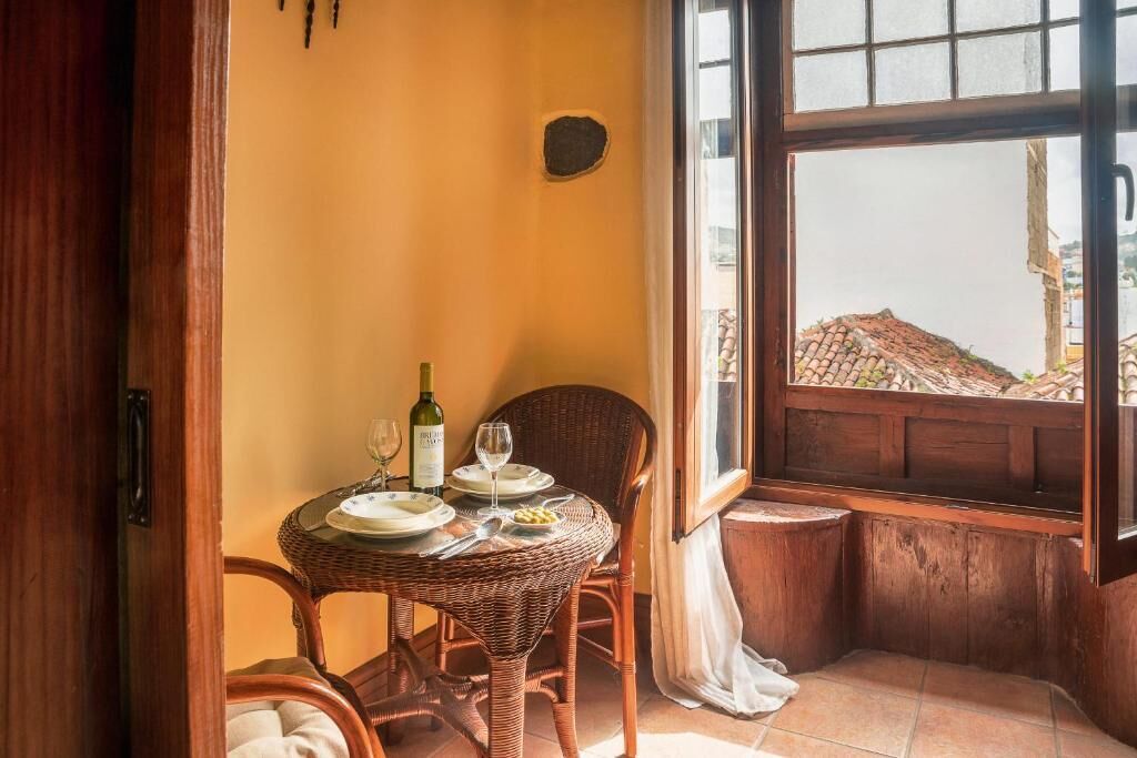Comedor con mesa de mimbre dispuesta para dos personas, una botella de vino y vistas a través de una ventana.