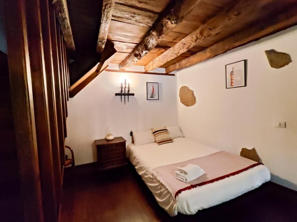 Ein Schlafzimmer mit Holzbalkendecke, einem Bett mit weißer und roter Decke, einem Nachttisch mit Lampe sowie einer Wand mit Bild und Kerzenhalter.