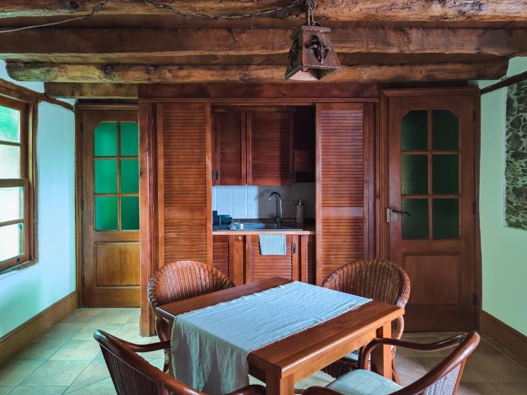 Een rustieke eethoek met een tafel en stoelen, een keuken met houten kasten, en een groene deur.
