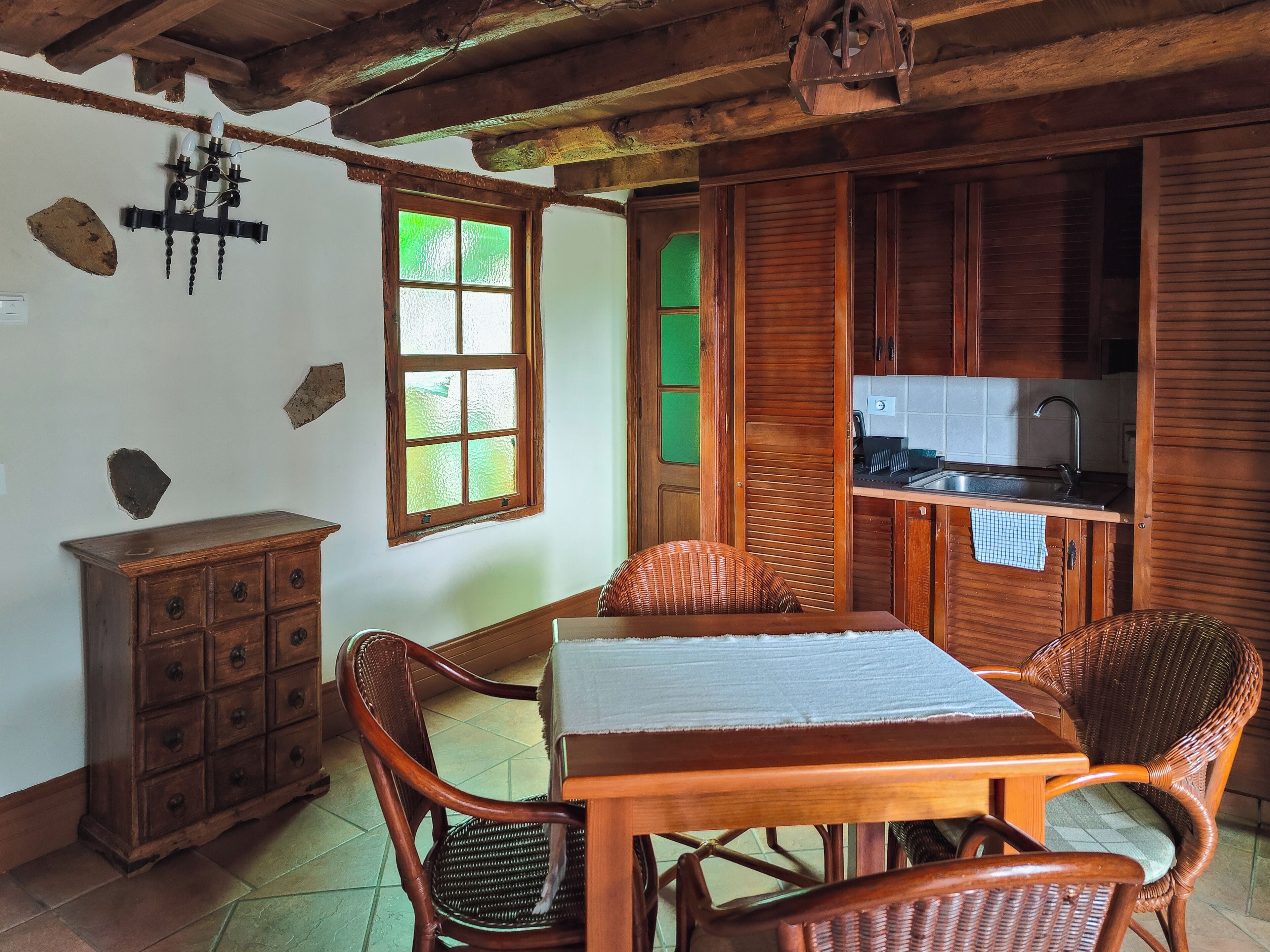 Una mesa de comedor de madera con sillas, un armario de madera, un ventanal con vidrio esmerilado y una zona de cocina con armarios de madera y un lavamanos.