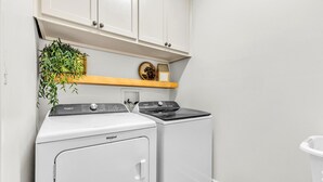A laundry room with a Whirlpool washer and dryer, a wooden shelf with decorative items, and a hanging plant. - Premier Lake Martin Lakefront Home with Private Boat Ramp, 38 Minutes to Auburn! (Dadeville)