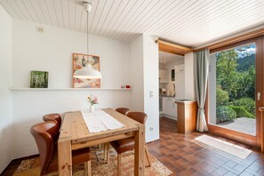 Ein Essbereich mit einem Holztisch, Stühlen, einer Hängelampe sowie Blick auf eine Küche und einen Garten durch eine Glastür.