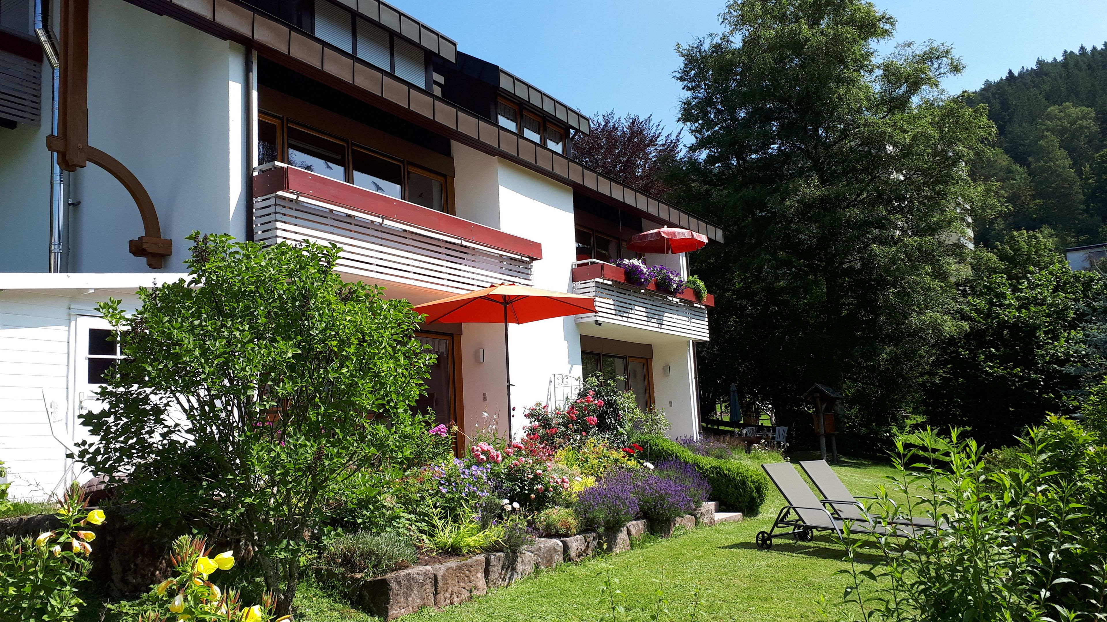 Een gebouw met twee verdiepingen, voorzien van een balkon, een tuin met diverse planten en bloemen, en een gazon met twee comfortabele stoelen.