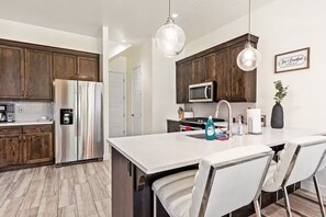 Una cocina moderna con armarios de madera, electrodomésticos de acero inoxidable, una isla blanca con taburetes y luminarias colgantes.