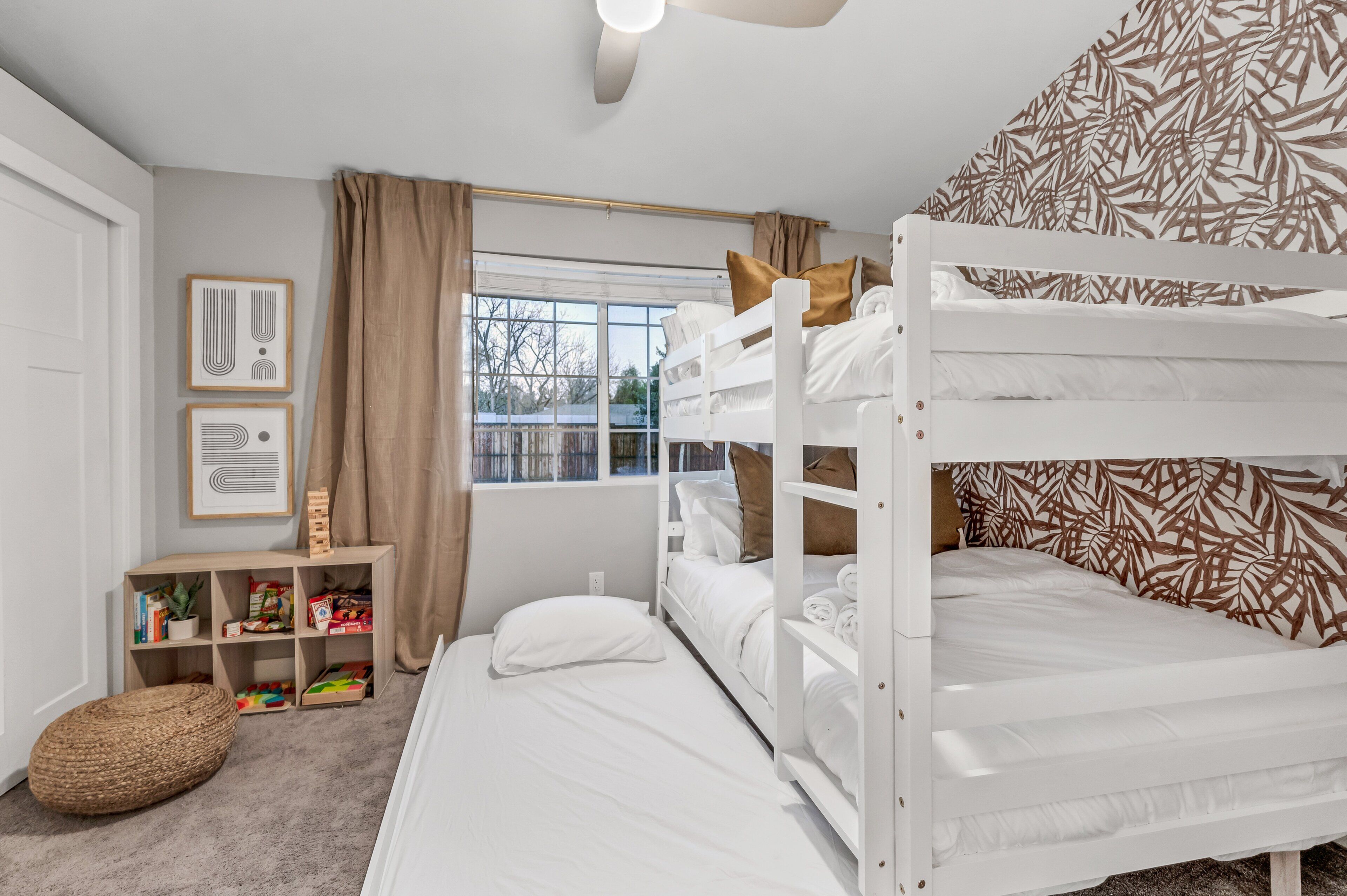 A room with bunk beds, a window with curtains, a bookshelf, and a wall with a leaf pattern.