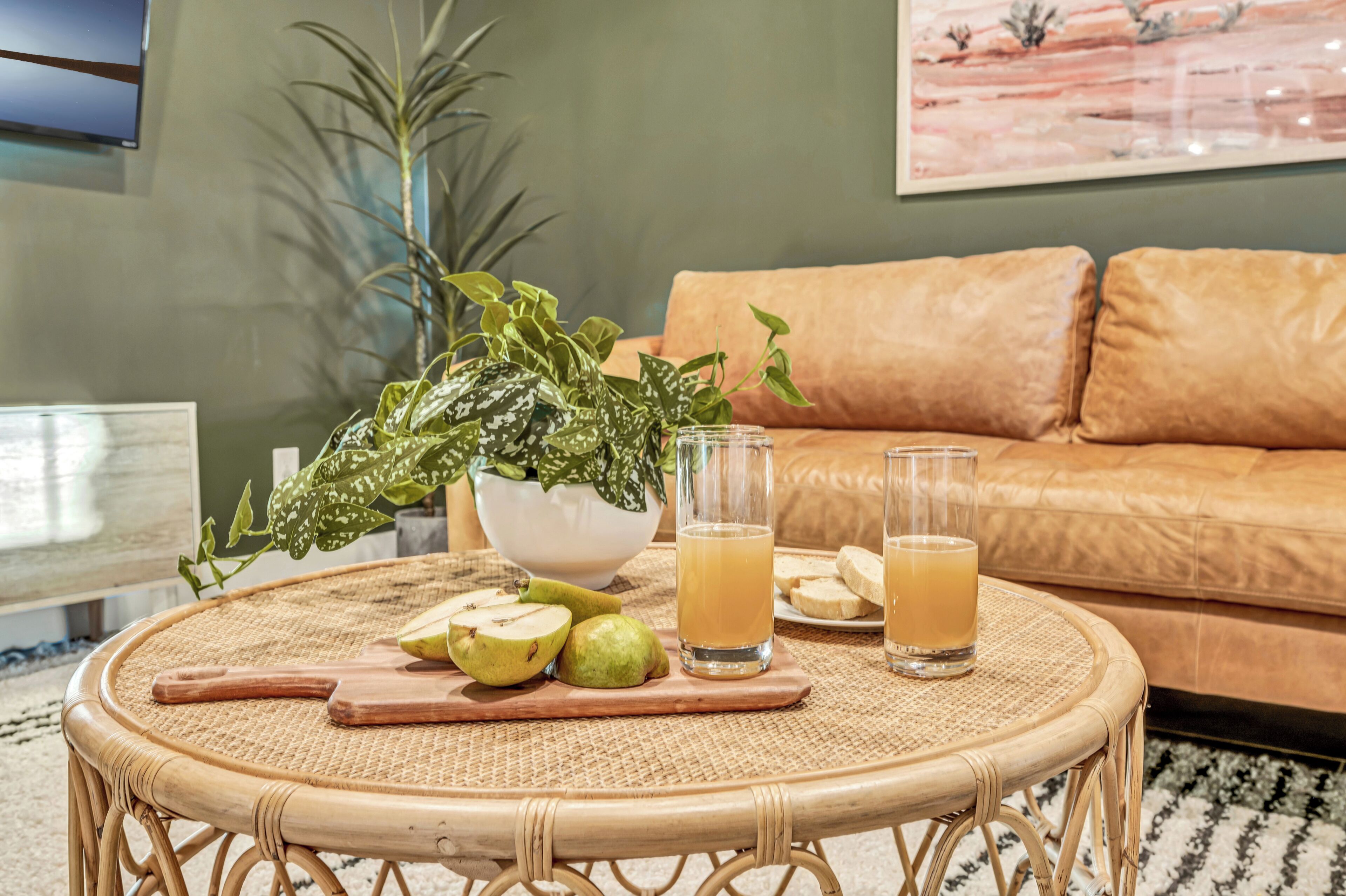 A round wicker table with a potted plant, a cutting board with fruit, and two glasses of juice.