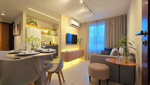 A modern living room with a sofa, ottoman, and a dining area with chairs and a table set for two. - Jardins do Atlântico By Hosppedar (João Pessoa)