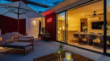 Un'area salotto esterna moderna con poltrone lounge e un tavolo, una porta in vetro che dà su uno spazio pranzo interno e un neon sulla parete.