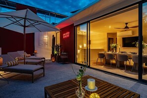 A modern outdoor seating area with lounge chairs and a table, a glass door leading to an interior dining space, and a neon sign on the wall.