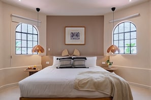 A bedroom with a large bed, two bedside lamps, a framed artwork above the bed, and a window with a view.
