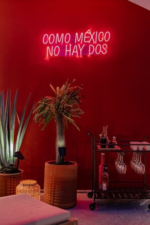 A room with a neon sign, a potted plant, a small table, and a cart with glasses and bottles.