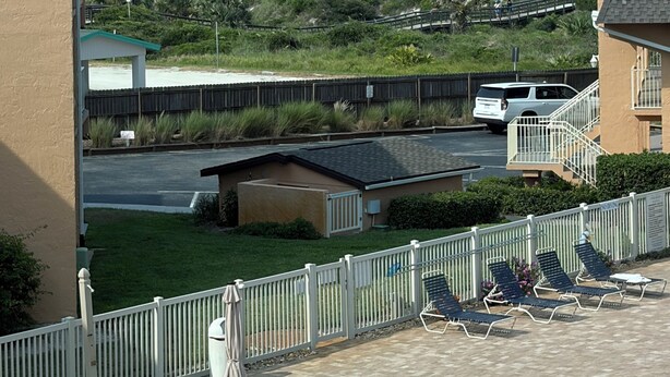 Un balcon avec vue sur l’océan, une terrasse clôturée avec des chaises longues et un espace de stationnement où se trouve un véhicule blanc.