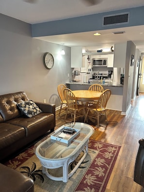 A living room with a brown leather sofa, a glass coffee table, and a dining area with a wooden table and chairs. - Beachfront condo with direct pool & ocean view on Crescent Beach (St. Augustine)