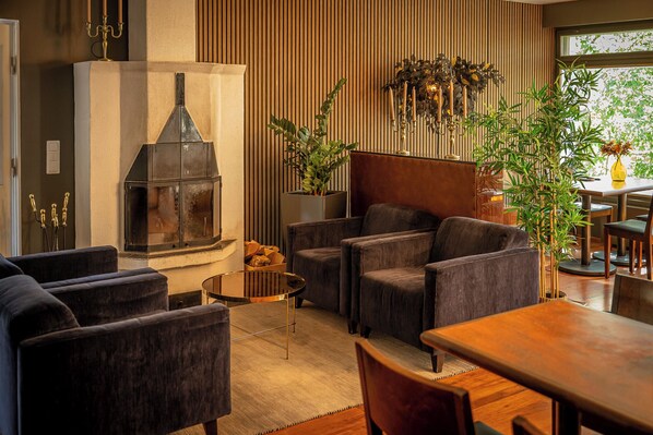 A modern living room with a fireplace, dark upholstered chairs, a wooden dining table, and a large window with plants.