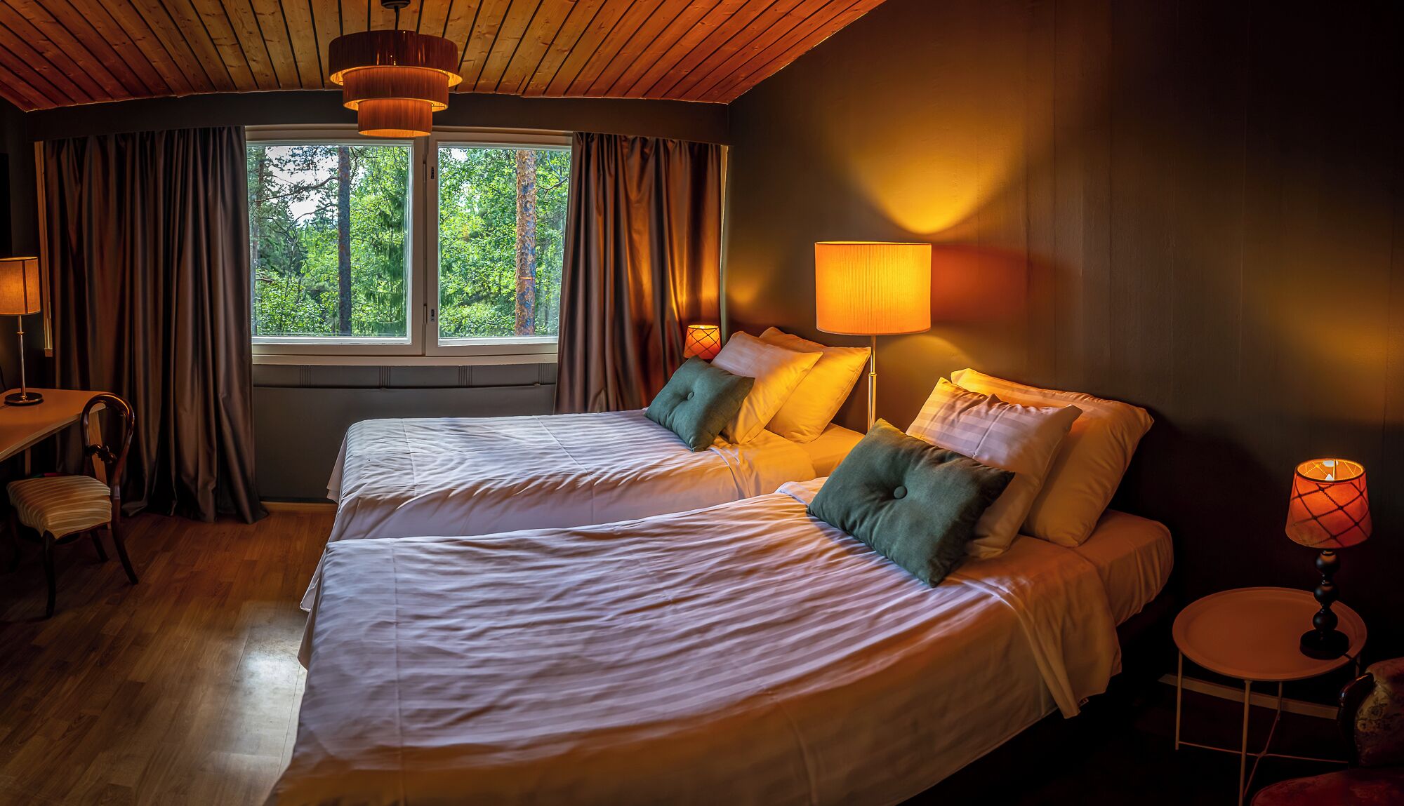 Een hotelkamer met twee bedden, een bureau en een stoel. Er is een raam met gordijnen, een lamp en uitzicht op bomen.