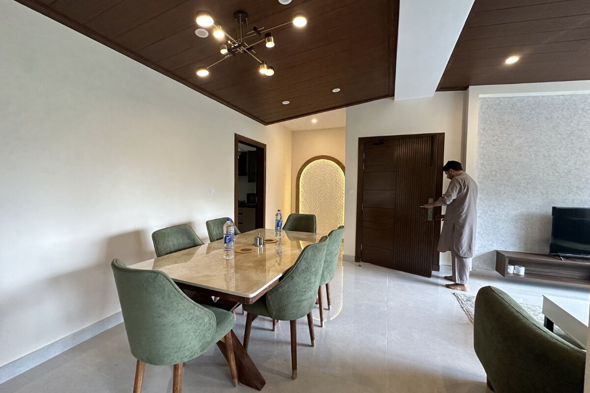 A modern dining area with a wooden table and green upholstered chairs, a person standing by a door, and a flat-screen TV on a stand.