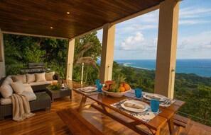 A dining area with a view of the ocean, a sofa, and a table set with plates and glasses. - Overhang Villa above ocean & Jungle-concierge (Bahía Ballena)