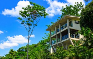 A multi-story building with balconies, surrounded by lush greenery and a clear blue sky. - Overhang Villa above ocean & Jungle-concierge (Bahía Ballena)