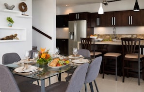 A modern kitchen with dark wooden cabinets, a stainless steel refrigerator, and a dining area with a glass table set for a meal. - Overhang Villa above ocean & Jungle-concierge (Bahía Ballena)