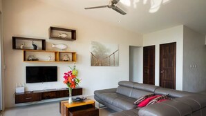 A modern living room with a grey sofa, a flat-screen TV, wooden shelves, and a painting of a bridge. - Luxury Designer villa Oceanview HR3 - Concierge (Bahía Ballena)