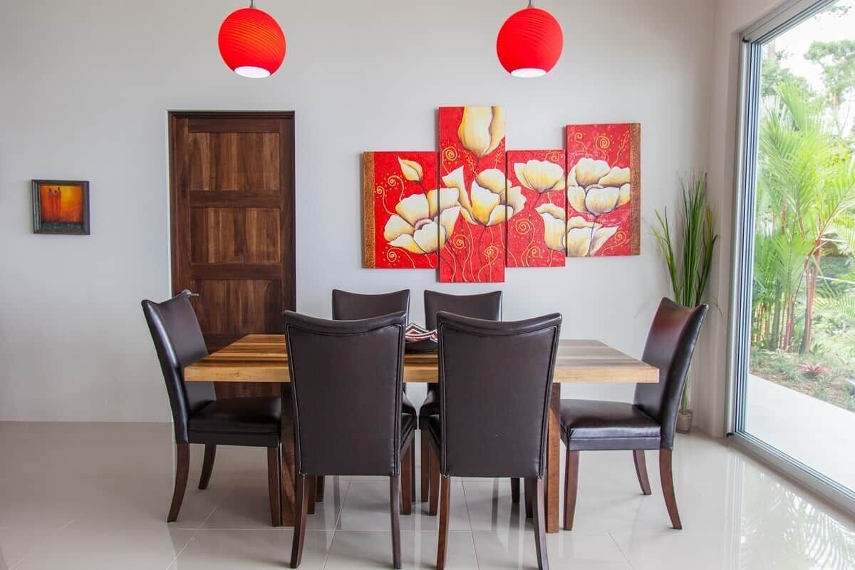 A dining area with a wooden table and chairs, a red door, and a large window with a view of greenery.