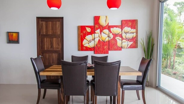 A dining area with a wooden table and chairs, a red door, and a large window with a view of greenery. - Luxury Designer villa Oceanview HR3 - Concierge (Bahía Ballena)