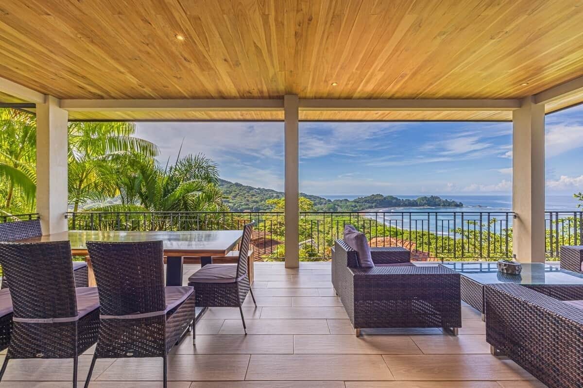An outdoor patio with wicker furniture, a glass table, and a view of the sea and hills.